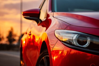 Crveni automobil snimljen u krupnom planu pri zalasku sunca, sa refleksijom neba i drveća na sjajnoj površini haube